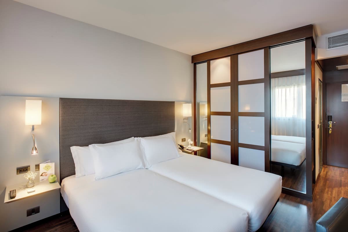 Ein Hotelzimmer mit einem großen Bett, einem Nachttisch mit Lampe, einem Spiegel und einer Holztür mit satinierten Glasscheiben.