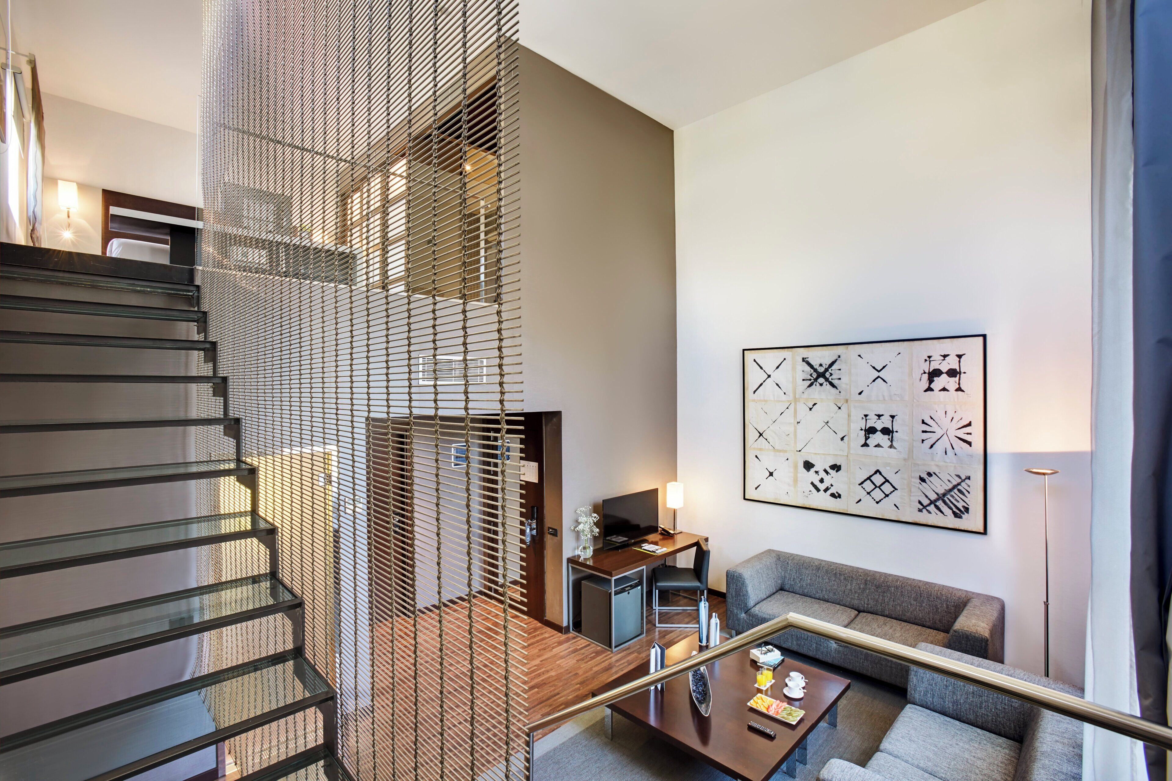 Una habitación de hotel moderna con una escalera, una zona de estar con sofá, un escritorio y una gran obra de arte abstracto en la pared.