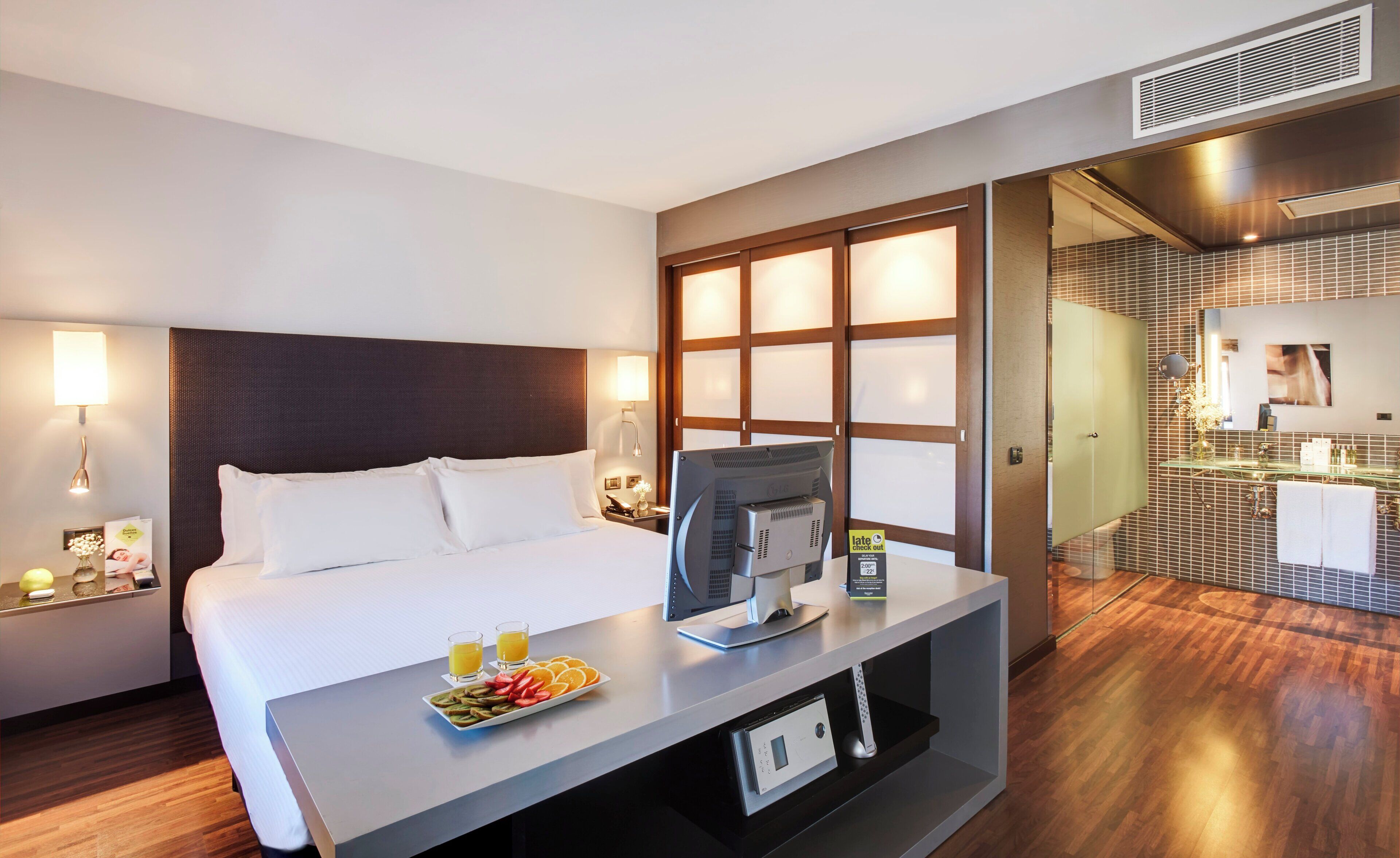 A hotel room with a bed, a desk with a computer, a tray of fruit, and a bathroom with a mirror and sink.