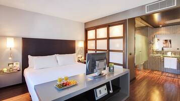 A hotel room with a bed, a desk with a computer, a tray of fruit, and a bathroom with a mirror and sink.