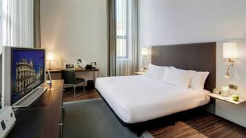 A hotel room with a large bed, a desk with a computer, a TV, and a window with curtains.
