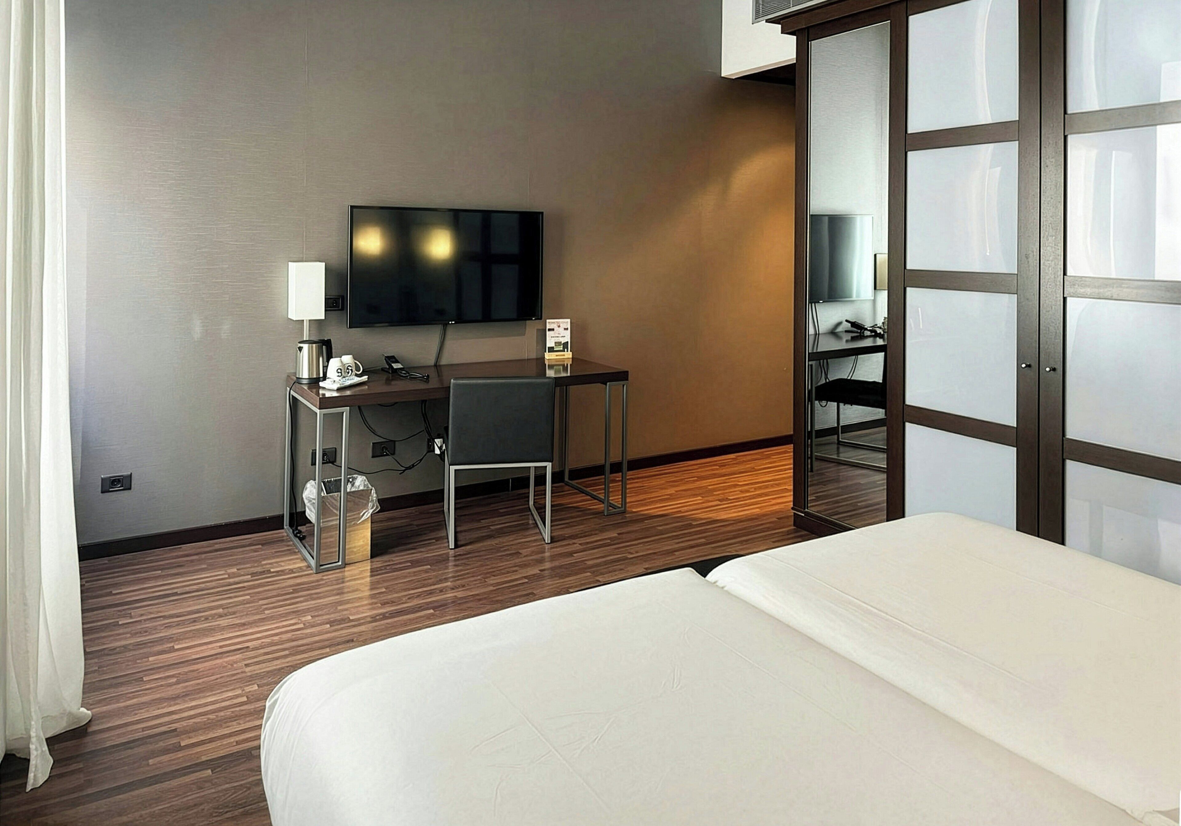 Habitación de hotel con cama, escritorio, computadora, televisión y ventana con paneles de vidrio esmerilado.