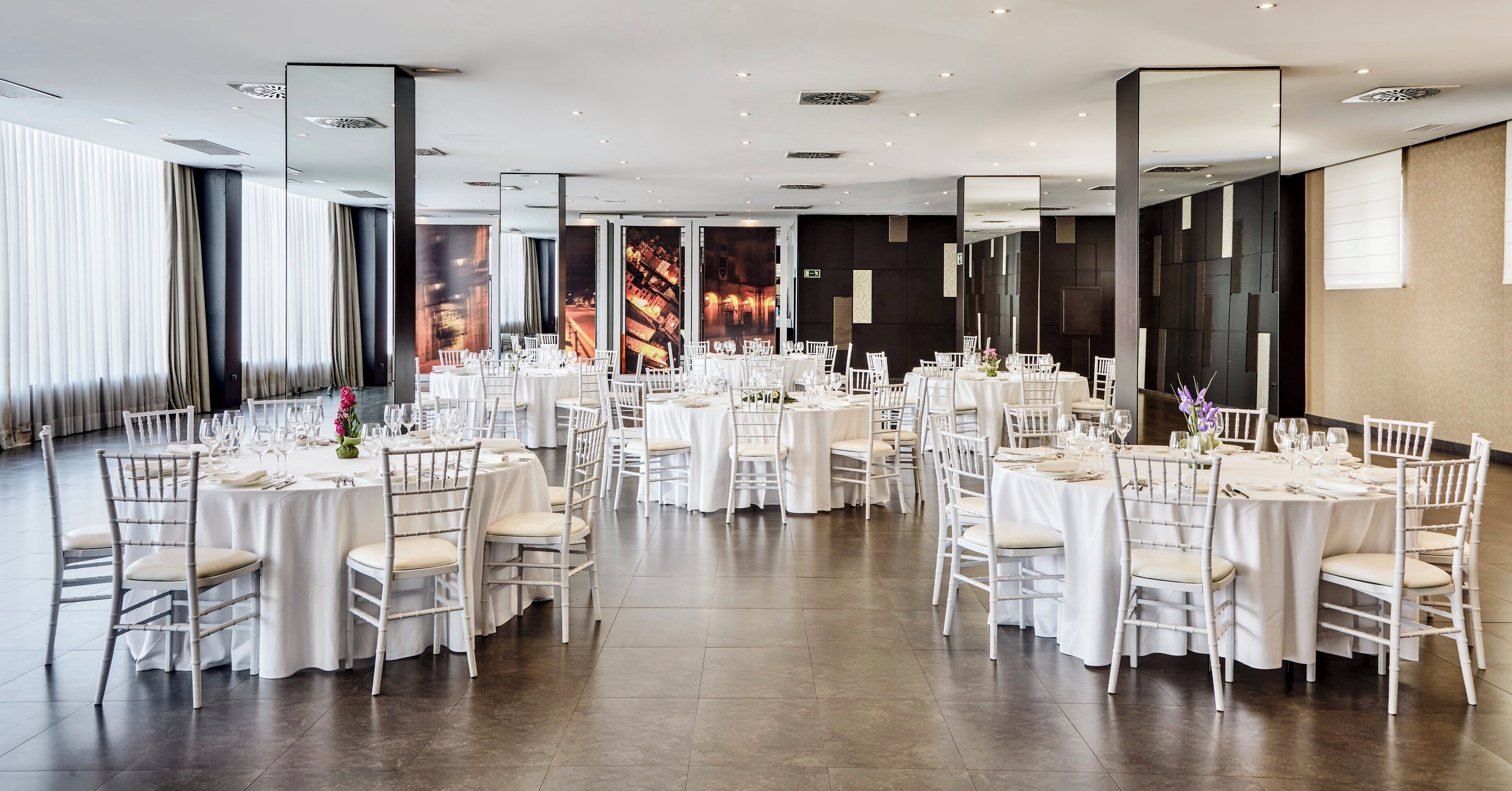A spacious banquet hall with round tables set for dining, white chairs, and a modern interior design.