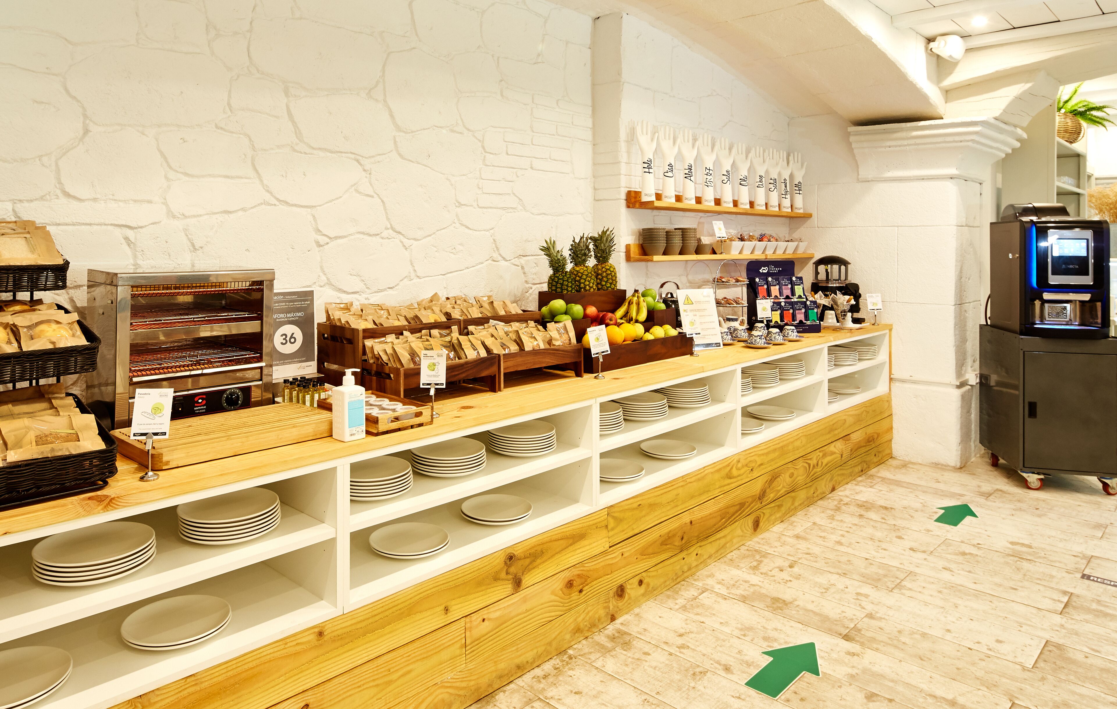 A bakery counter with bread, pastries, and a coffee machine. There are shelves with plates and a display of fruits and vegetables.
