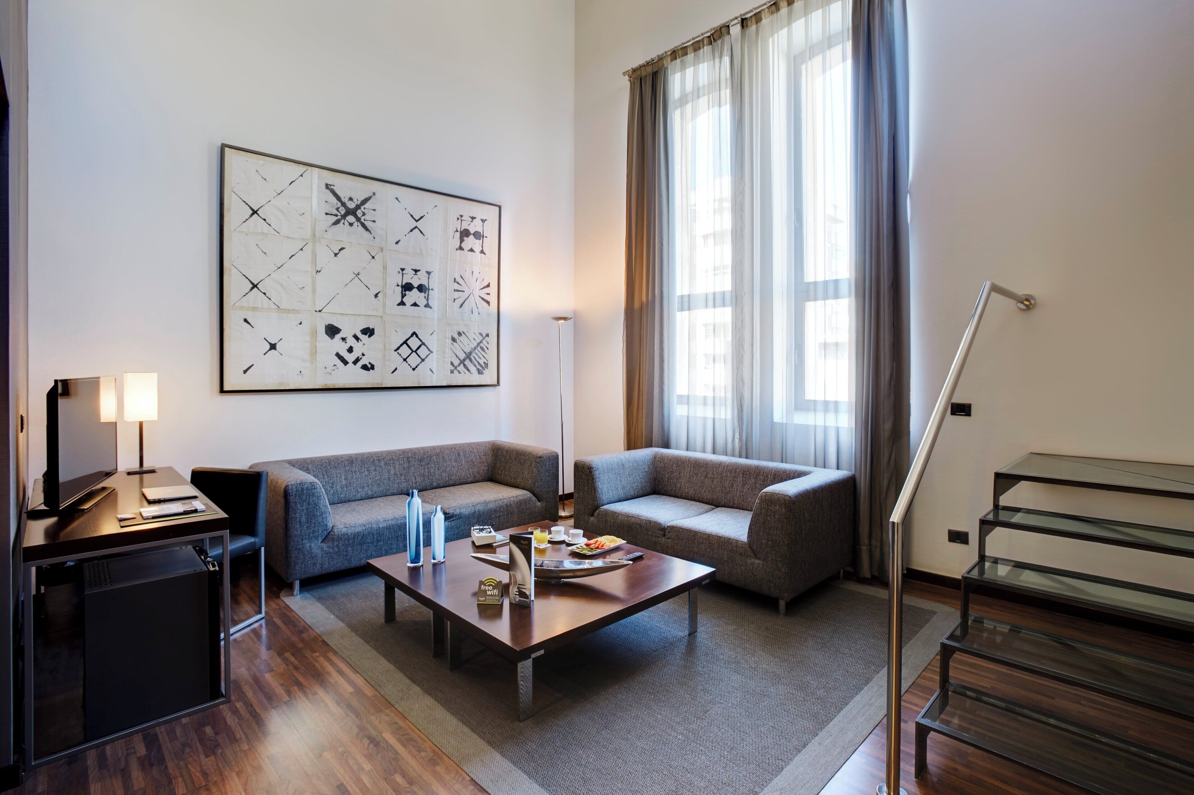 Una habitación de hotel moderna con un conjunto de sofás, una mesa de centro con alimentos para el desayuno, un escritorio con lámpara y un cuadro abstracto grande en la pared.