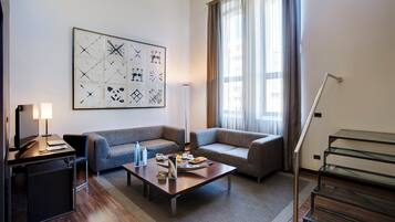 Una habitación de hotel moderna con un conjunto de sofás, una mesa de centro con alimentos para el desayuno, un escritorio con lámpara y un cuadro abstracto grande en la pared.