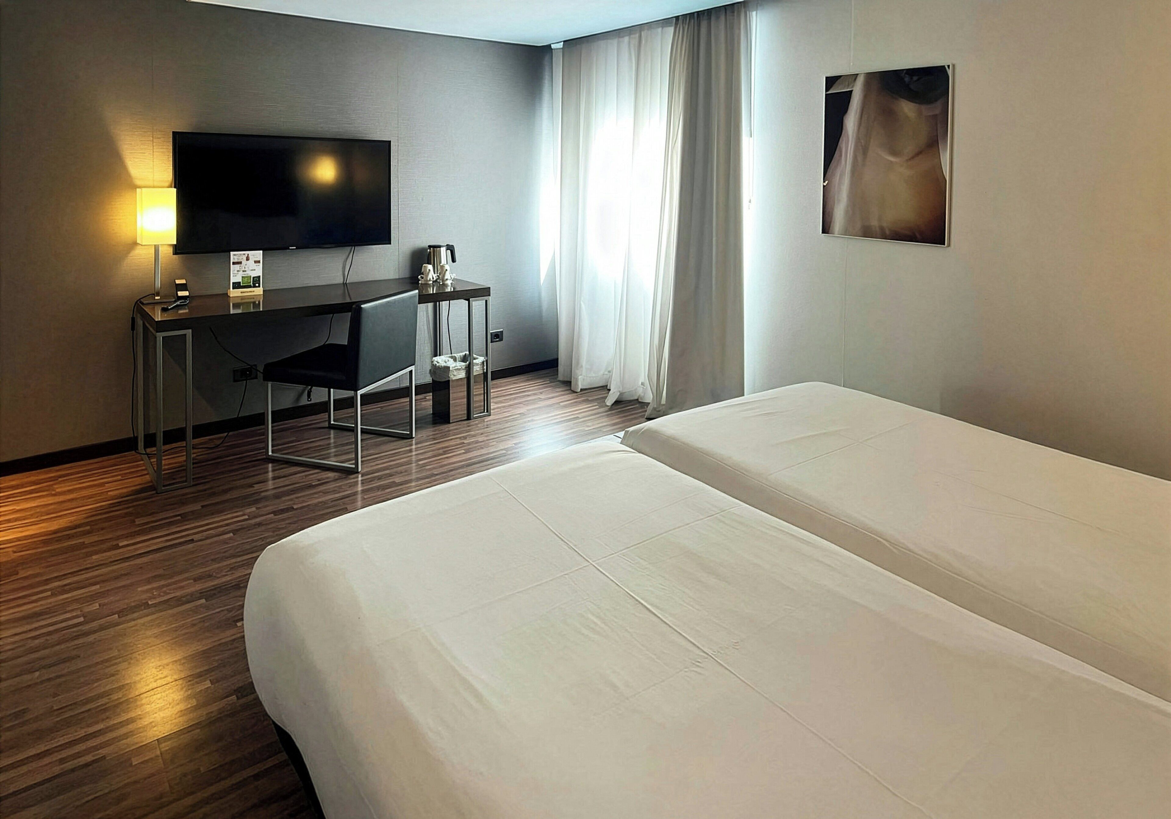 Habitación de hotel con cama, escritorio con televisor, cafetera y ventana con cortinas.