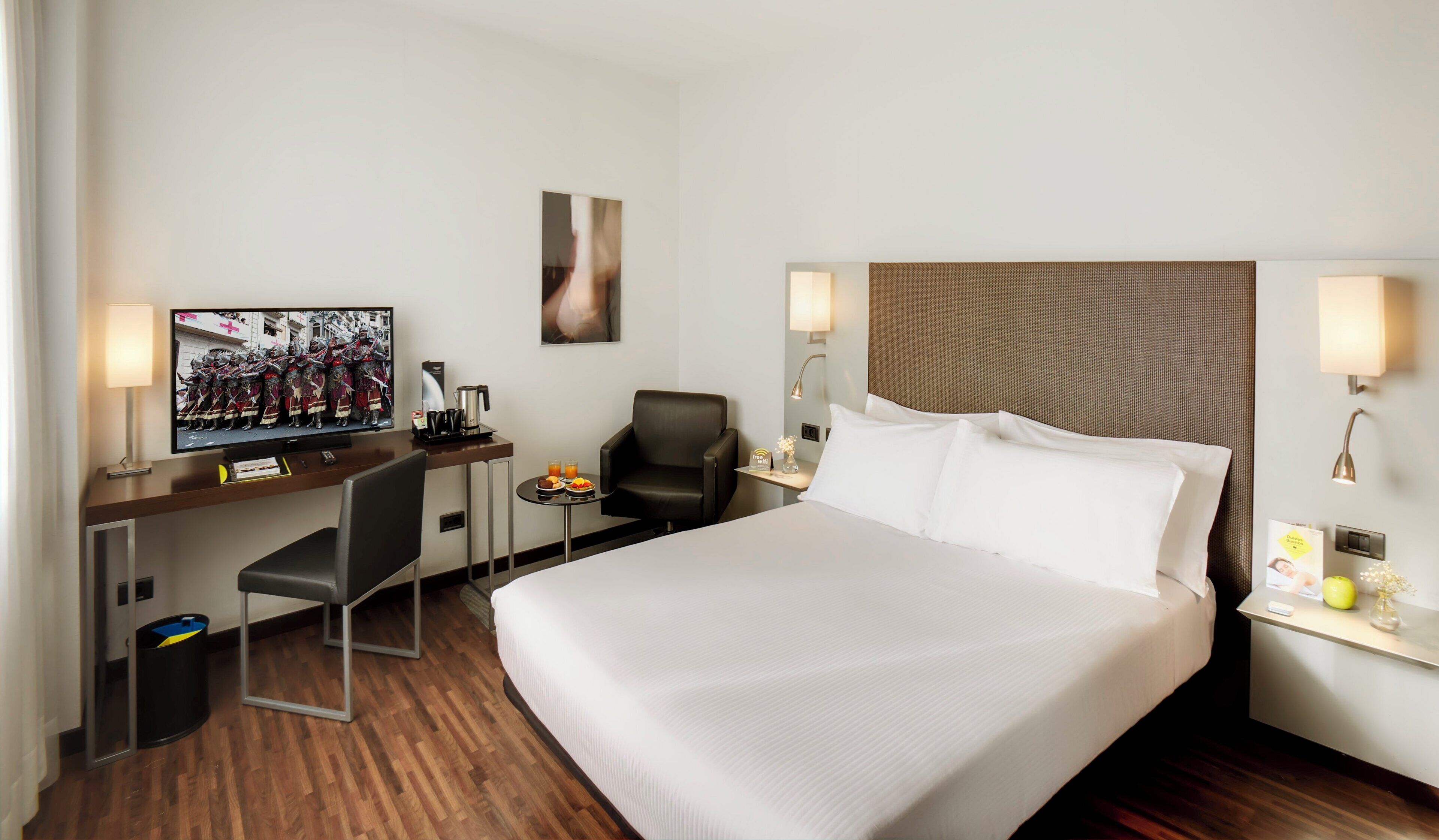 A hotel room with a bed, a desk with a television, a chair, a small table with fruit, and a trash bin.