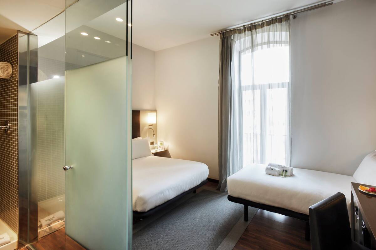 Ein Hotelzimmer mit zwei Betten, einem Schreibtisch mit einem Laptop, einer Duschkabine und einem großen Fenster mit Vorhängen.