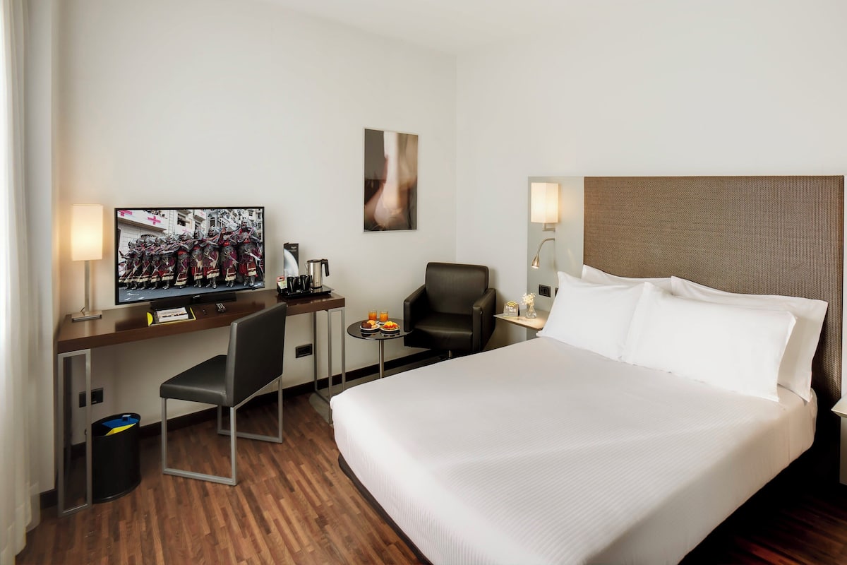 Ein Hotelzimmer mit einem Bett, einem Schreibtisch mit Fernseher, einem Stuhl, einem kleinen Tisch mit Früchten und einem Mülleimer.