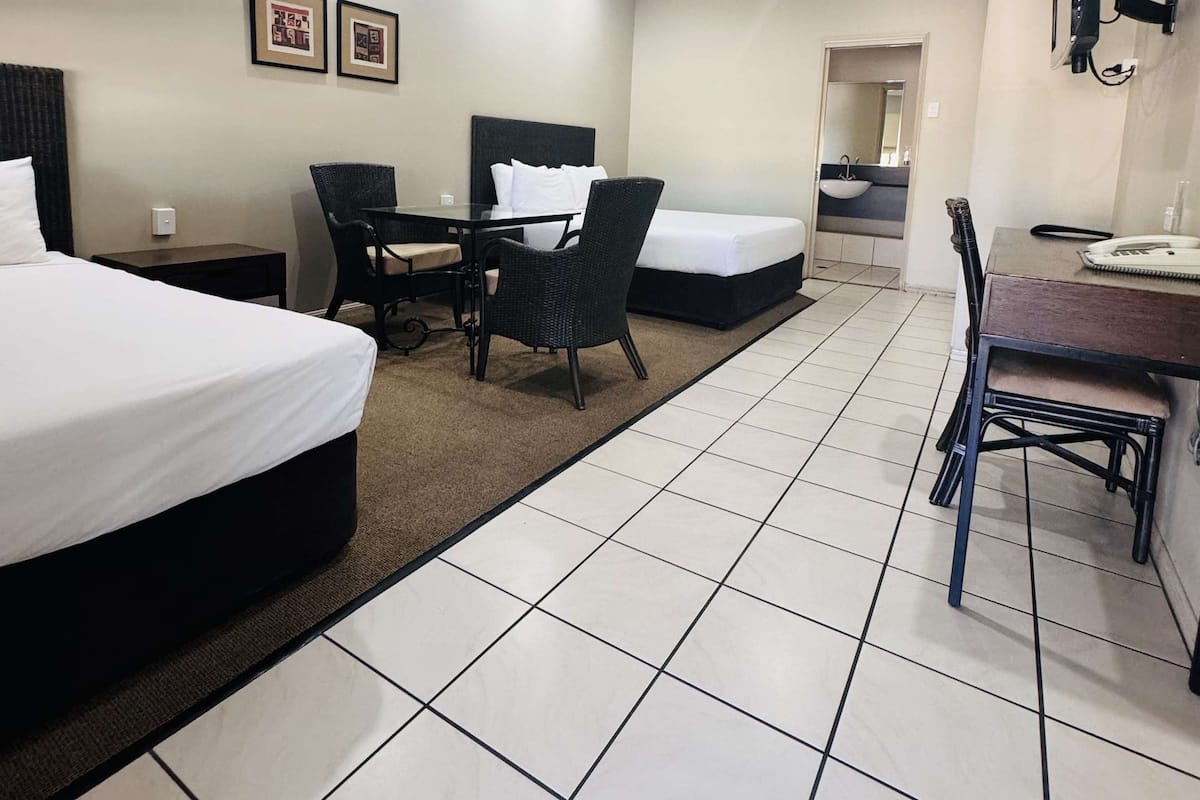 A hotel room with two beds, a desk, a chair, a small table, and a bathroom visible through an open door.