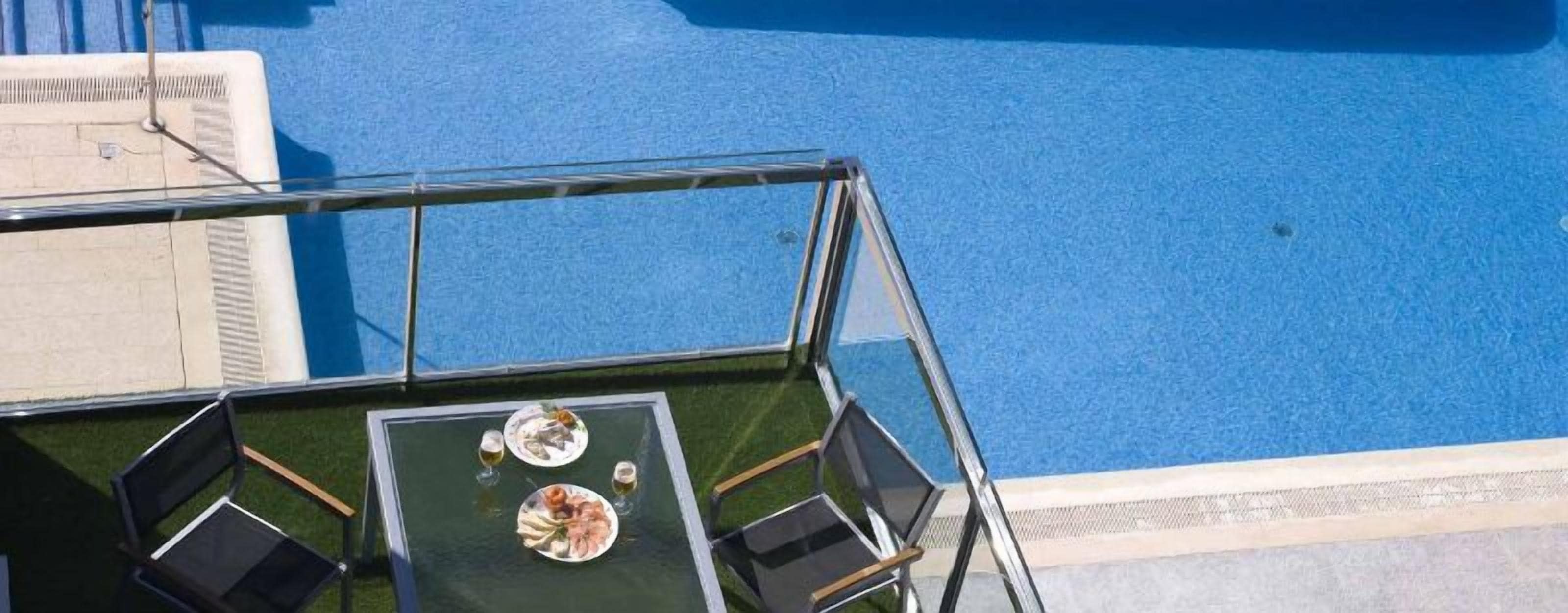 A rooftop pool area with glass railings, two tables with chairs, and a clear view of the pool.