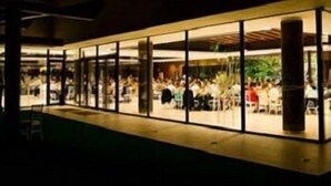 A modern building with a glass facade, a covered outdoor seating area, and a visible interior space with tables and chairs.