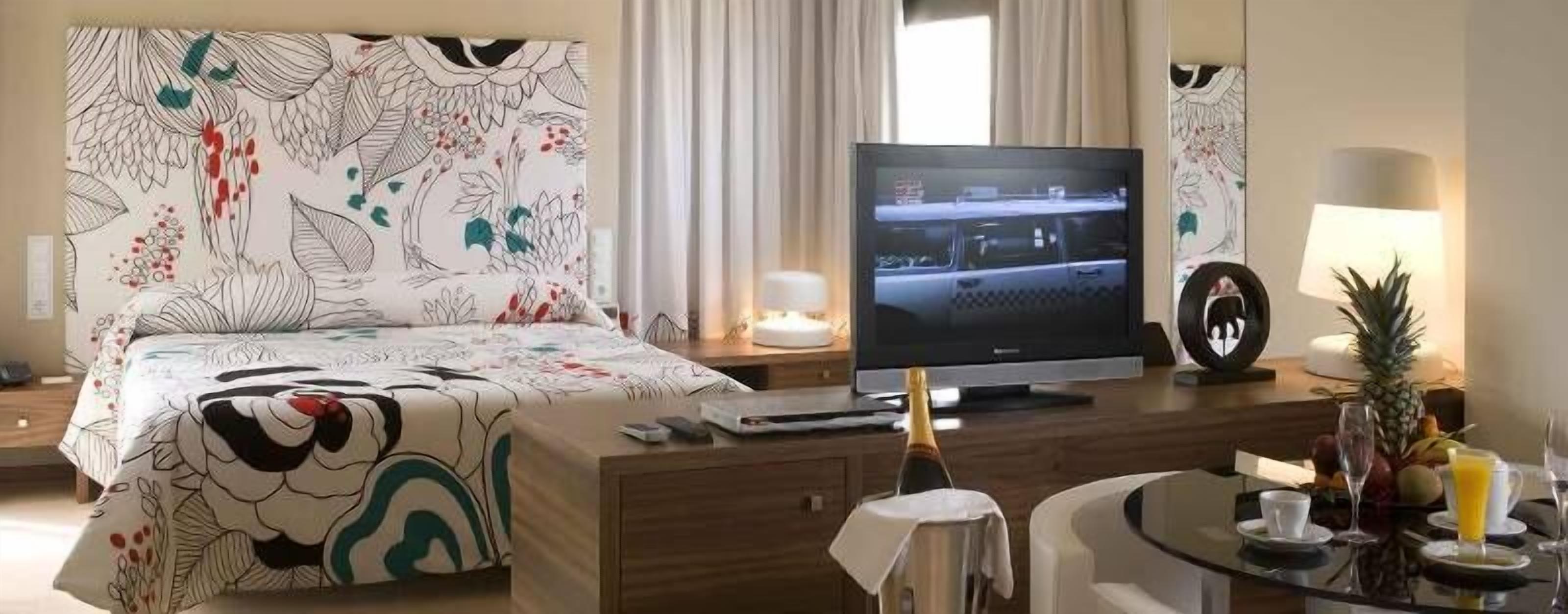 A hotel room with a bed, a TV, a desk with a lamp, and a dining area with fruit and drinks.