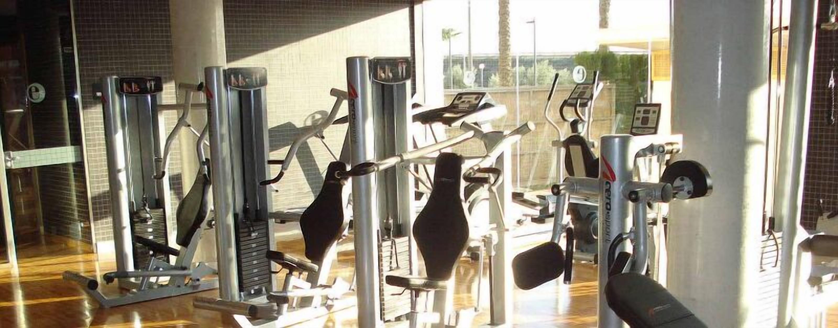 Een sportschool met diverse fitnessapparaten, een bakstenen muur en een raam met uitzicht op palmbomen.