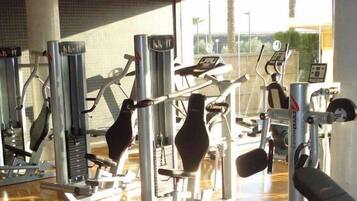 Een sportschool met diverse fitnessapparaten, een bakstenen muur en een raam met uitzicht op palmbomen.