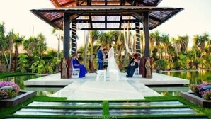 A gazebo with a thatched roof, surrounded by a garden with palm trees and a pond.