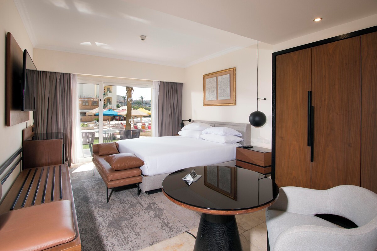 A hotel room with a bed, a desk, a chair, a TV, and a view of the outside.