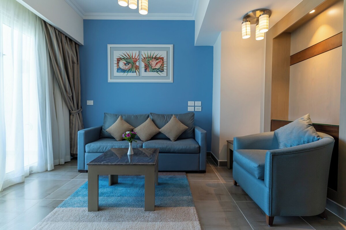 A modern living room with a blue sofa, a matching armchair, and a coffee table. There is a framed artwork on the wall and a window with curtains.
