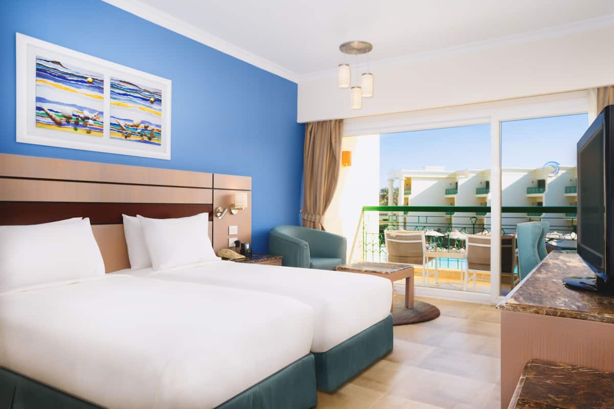 A hotel room with a large bed, a TV, a balcony with a view of buildings, and a painting on the wall.