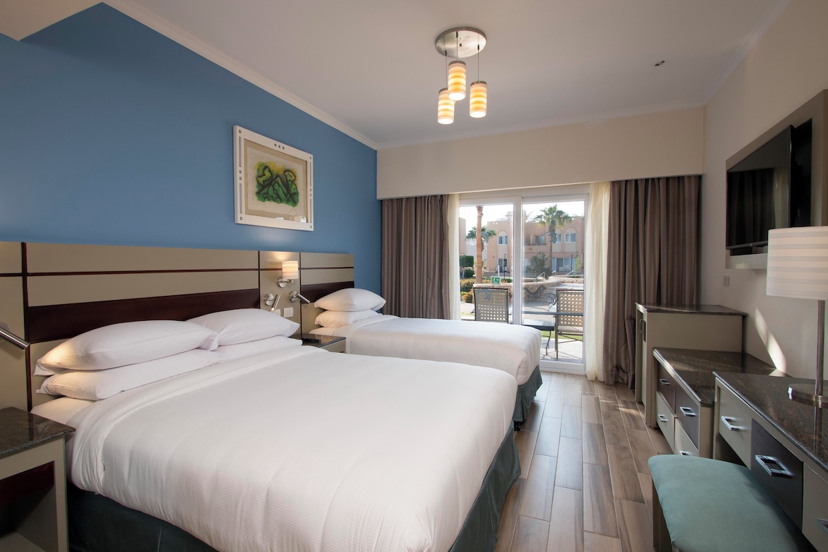 A hotel room with two beds, a painting on the wall, a TV, and a balcony with curtains.