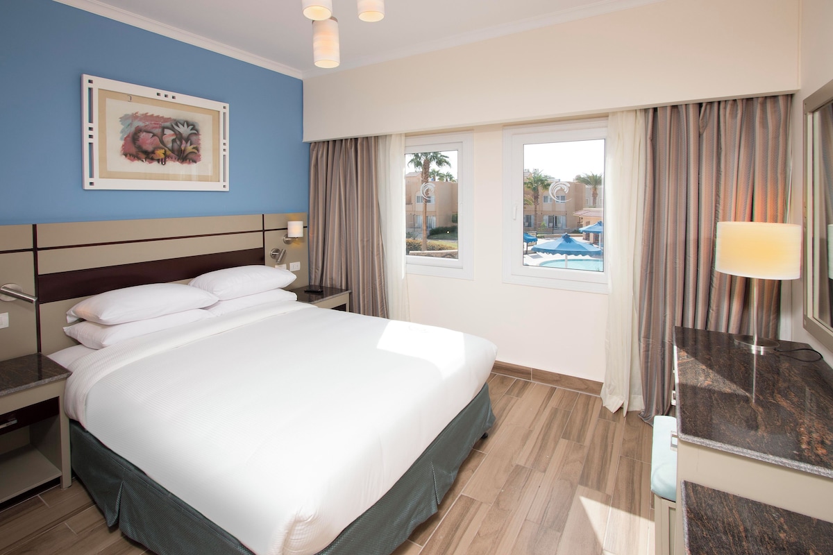 A hotel room with a large bed, a nightstand, a desk, a window with curtains, and a view of a swimming pool.