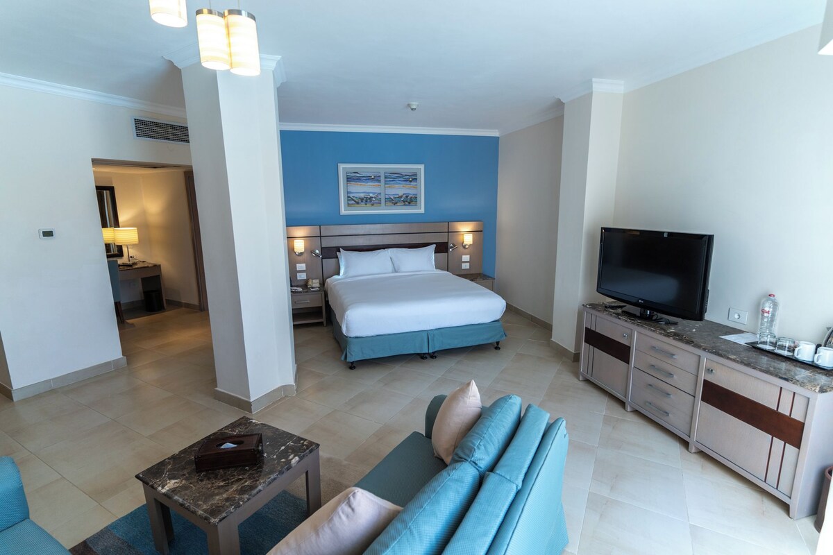 A hotel room with a bed, a TV, a sofa, a desk, and a bathroom visible in the background.