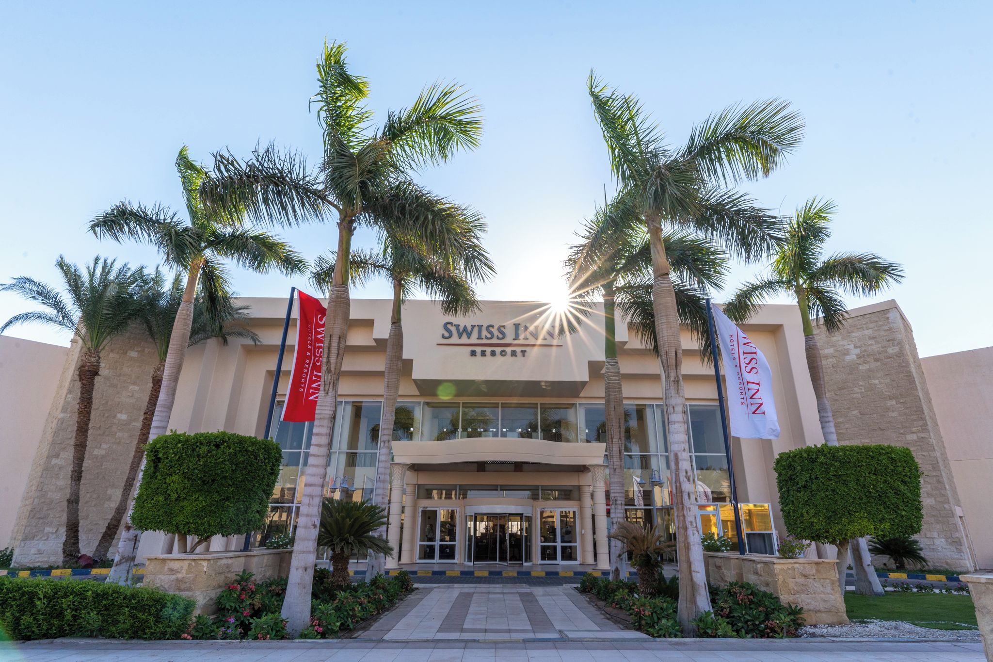 A hotel entrance with palm trees, a sign for 'SWISS INN RESORT', and flags.