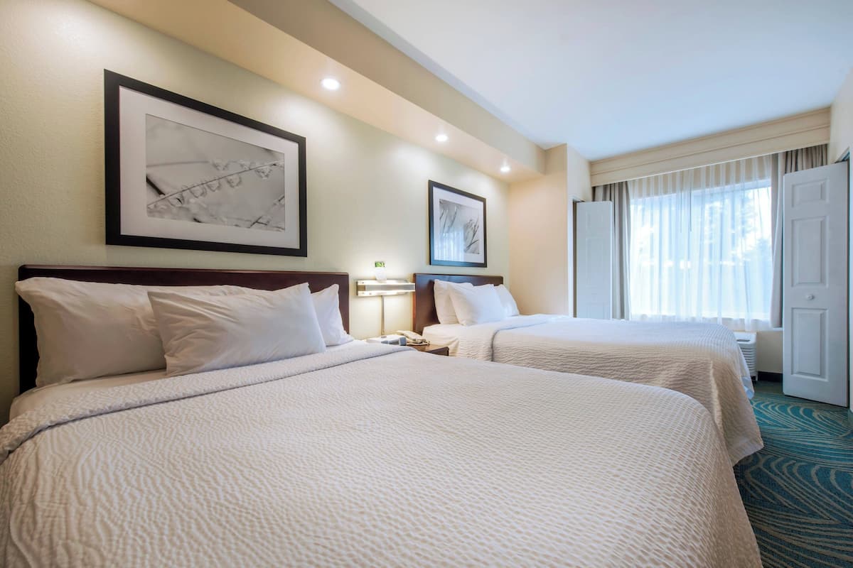 A hotel room with two beds, a large framed picture on the wall, a nightstand with a lamp, and a window with sheer curtains.
