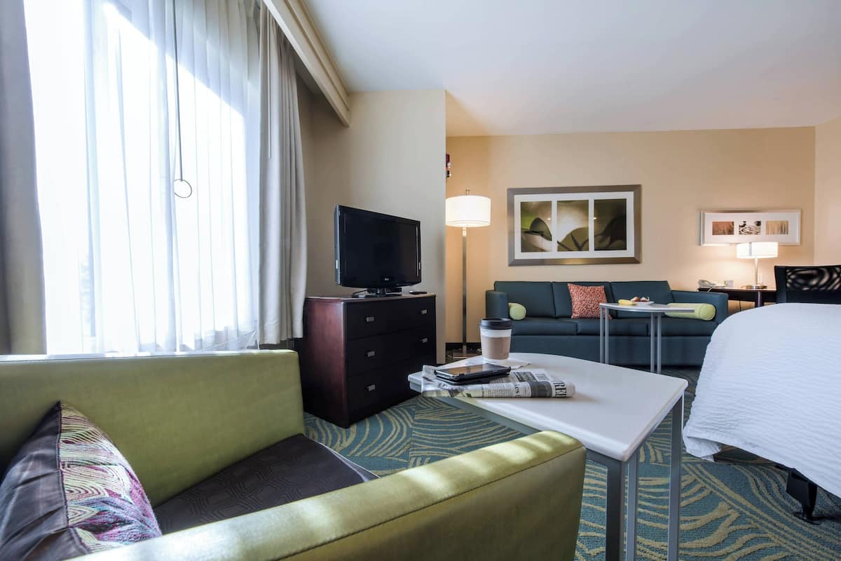 A hotel room with a bed, a sofa, a coffee table, a TV on a dresser, and a lamp.