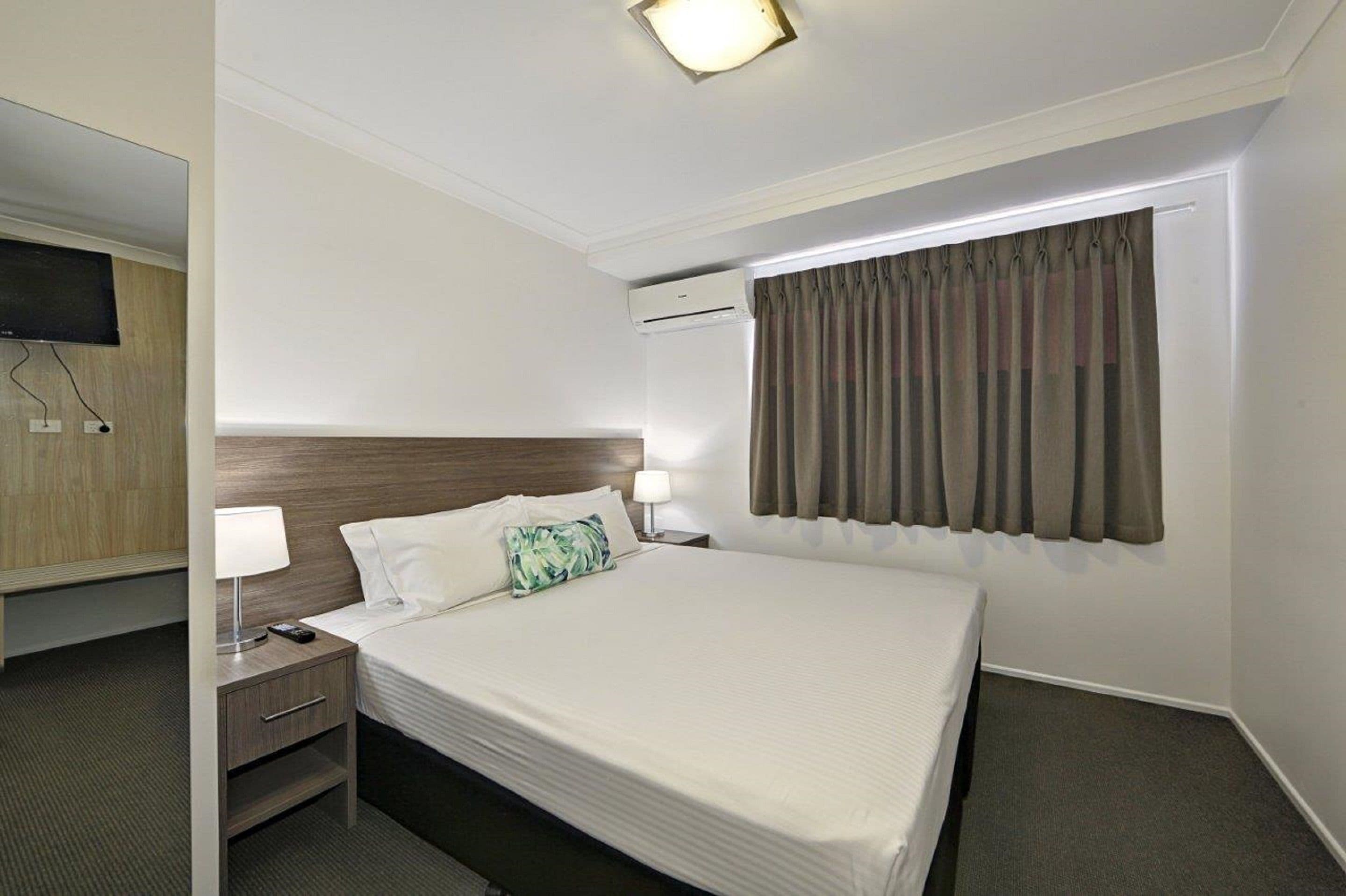 Habitación de hotel con una cama bien hecha, una lámpara de noche, un televisor de pantalla plana montado en la pared y un equipo de aire acondicionado.