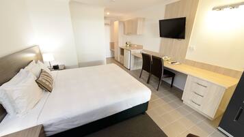 Quarto de hotel com cama, escrivaninha, cadeira, televisão e cozinha compacta.