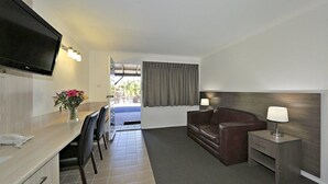 Quarto de hotel com televisão de tela plana, uma escrivaninha com telefone, um sofá de couro marrom, uma mesinha de cabeceira com abajur e vista para o exterior através de uma porta.
