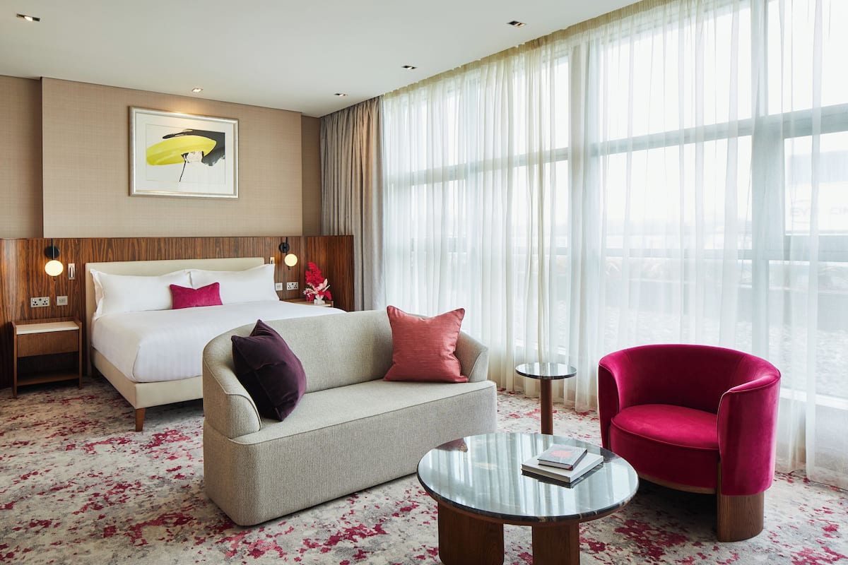 Hotelkamer met een bed, een slaapbank, een rode fauteuil, een kleine tafel en een groot raam met dunne gordijnen.