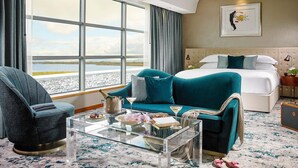 A hotel room with a large window, a bed, a teal sofa, a glass coffee table, and a chair.