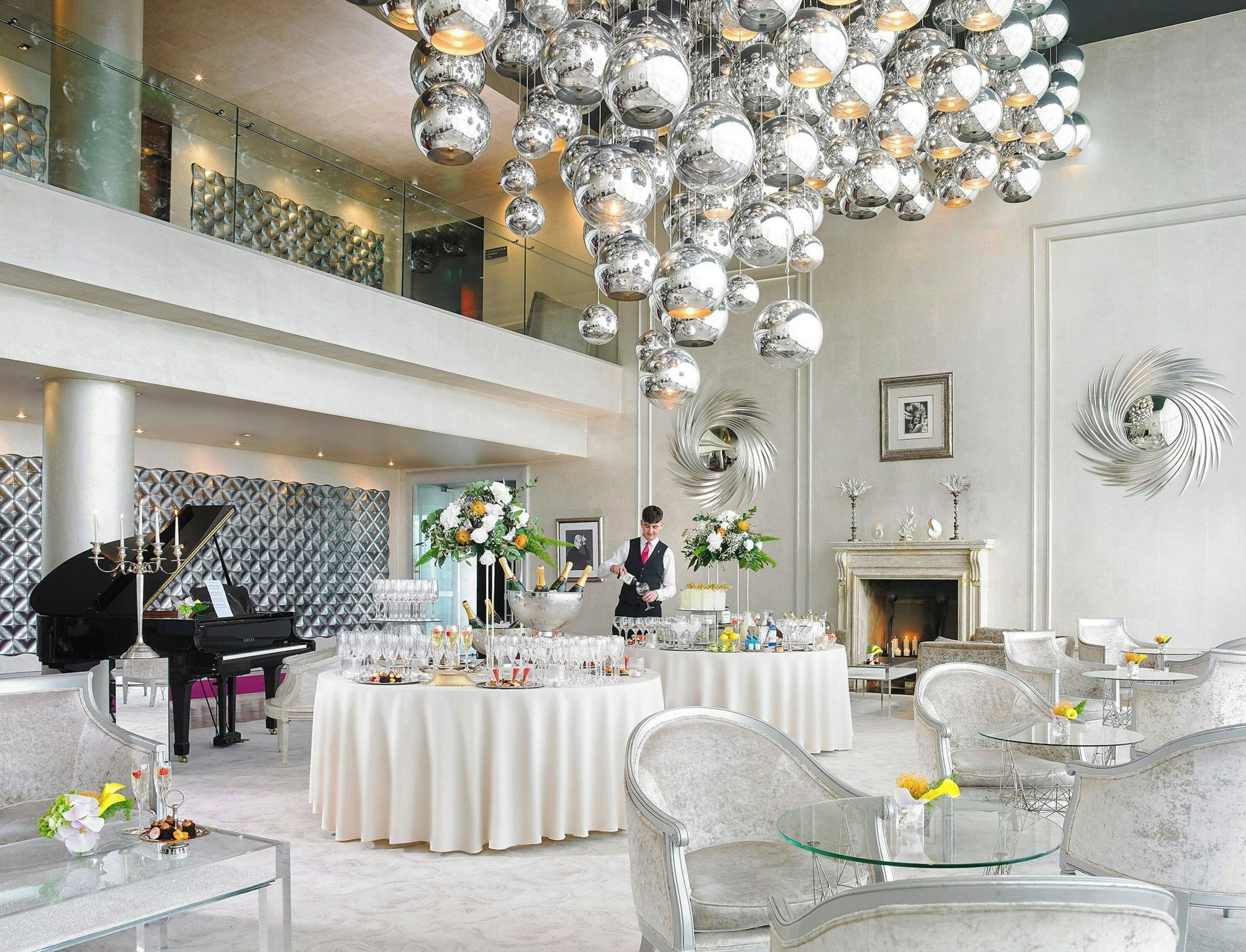 A spacious, elegantly decorated hotel lobby with a grand piano, multiple round tables set for dining, and a high ceiling with reflective spherical light fixtures.