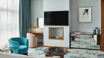 A modern hotel room with a blue armchair, a round coffee table, a flat-screen TV mounted on the wall, a small side table, and a framed artwork above the coffee table.