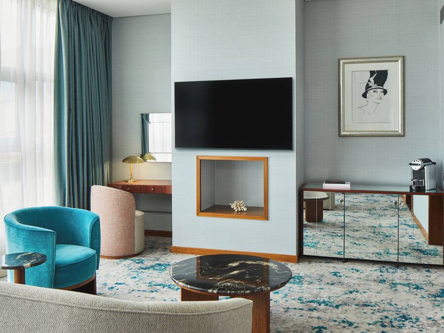 A modern hotel room with a blue armchair, a round coffee table, a flat-screen TV mounted on the wall, a small side table, and a framed artwork above the coffee table.