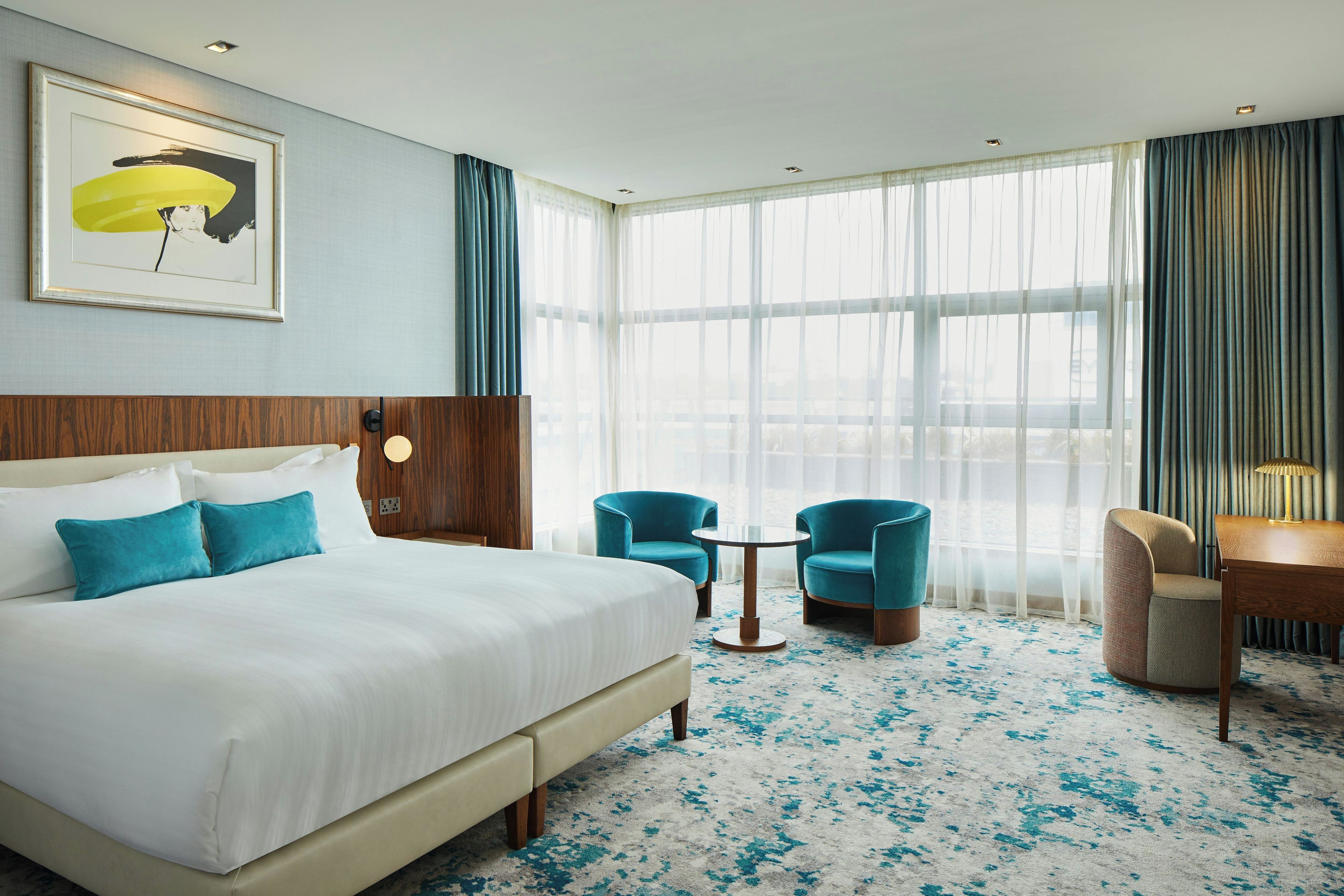 A hotel room with a large bed, two armchairs, a small table, and a view of the city through sheer curtains.