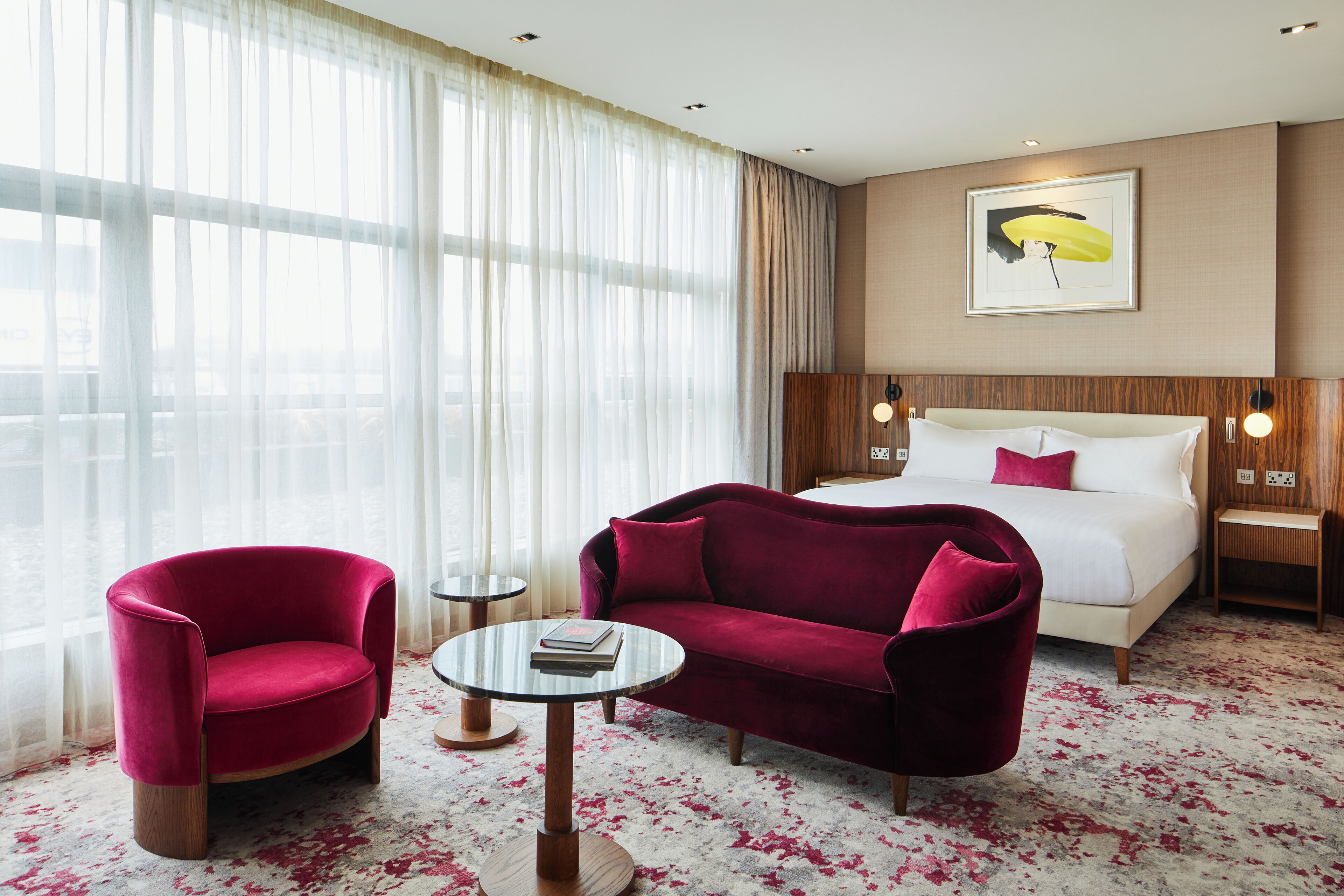A hotel room with a bed, a red armchair, a red sofa, a small round table, and a large window with sheer curtains.