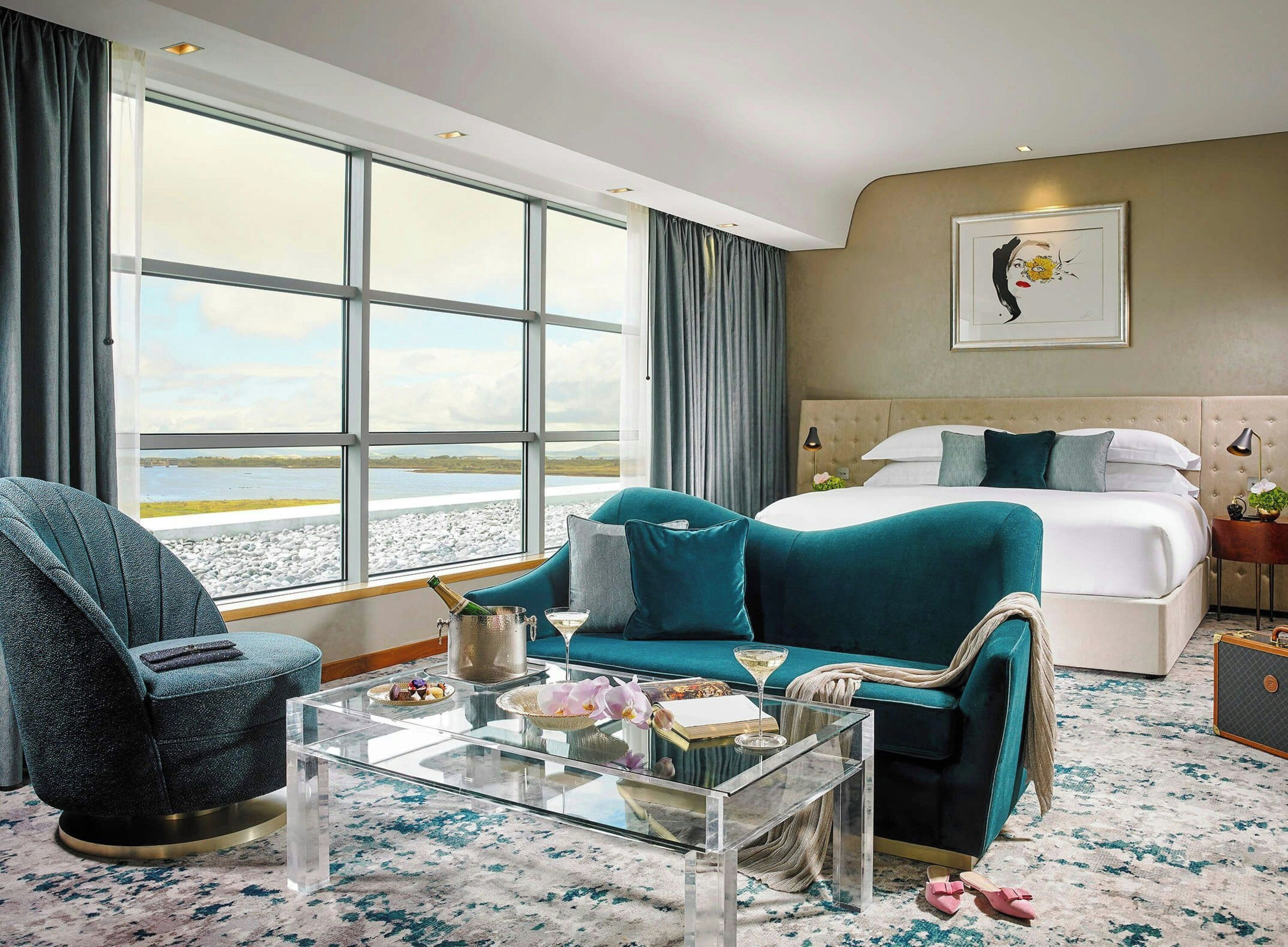 A hotel room with a large window, a bed, a sofa, a glass coffee table, and a chair.