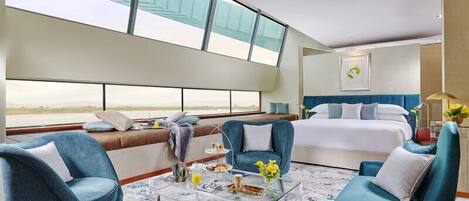 Ein modernes Hotelzimmer mit einem großen Bett, zwei blauen Sesseln, einem Glastisch und Meerblick durch große Fenster.