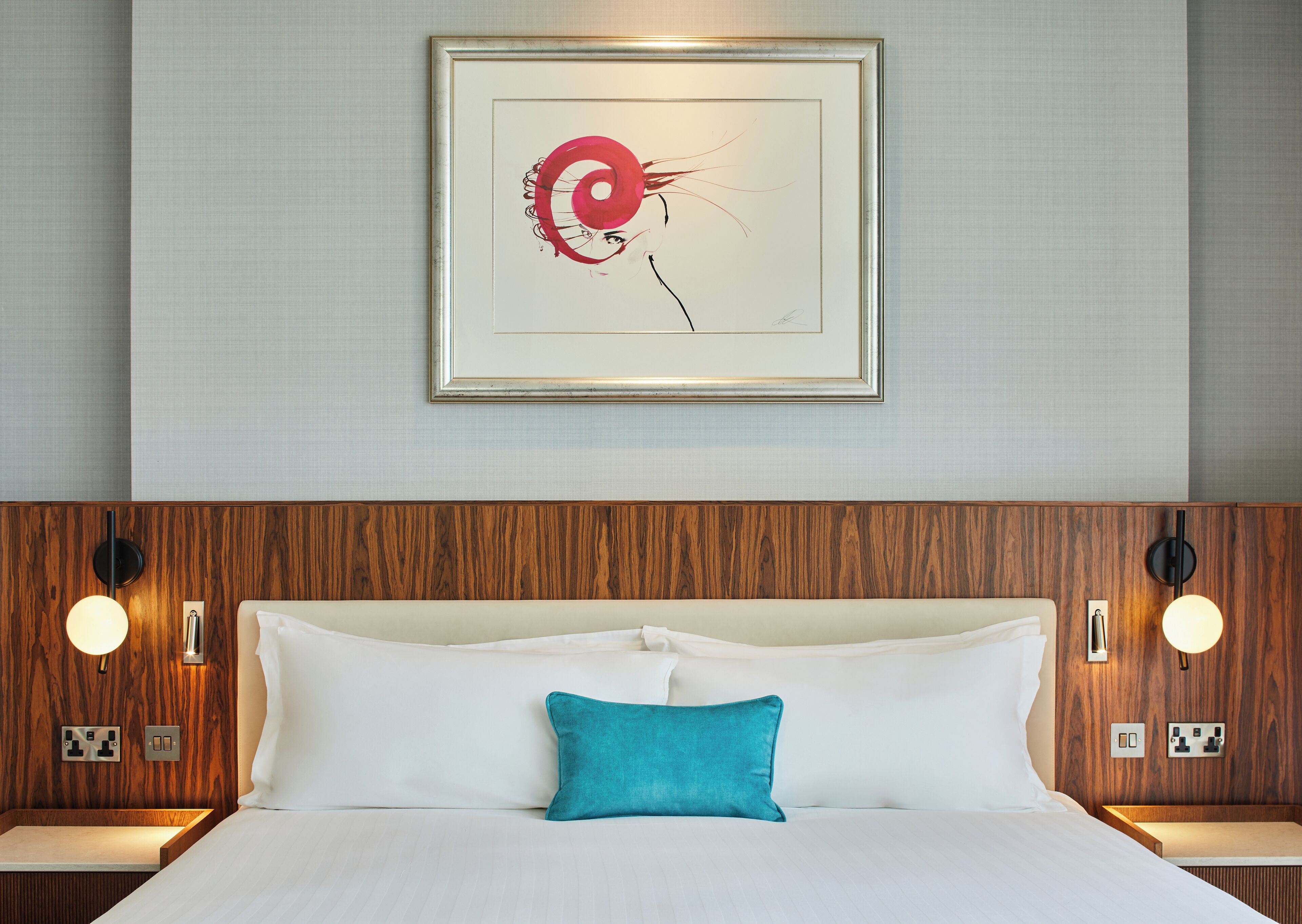 Una habitación de hotel con cabecera de madera, cama blanca con almohada verde azulada y una obra enmarcada sobre la cama.