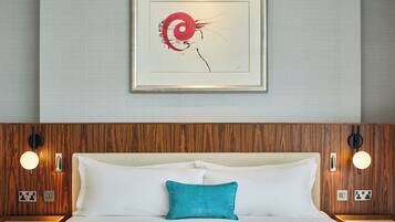 Una habitación de hotel con cabecera de madera, cama blanca con almohada verde azulada y una obra enmarcada sobre la cama.
