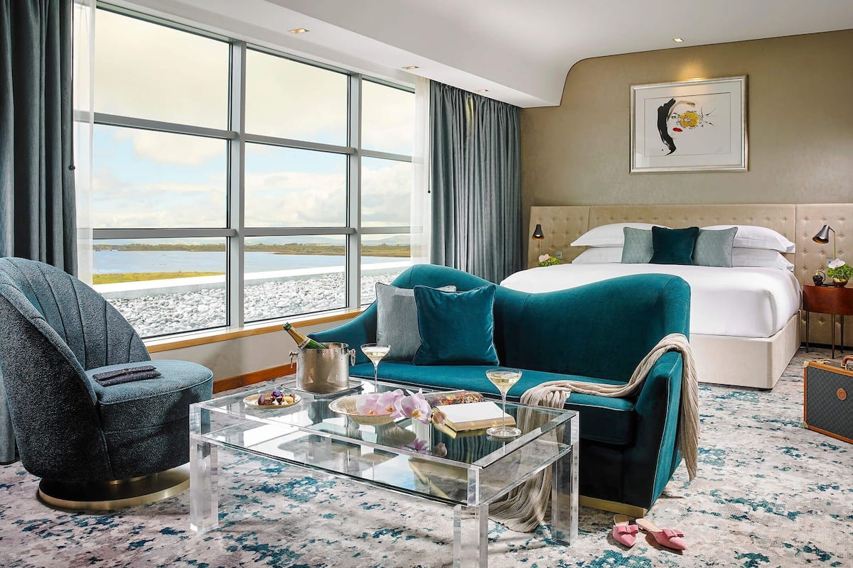 Hotelkamer met een groot raam, een bed, een blauwgroene bank, een glazen salontafel en uitzicht op het strand.