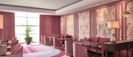A hotel lobby with a modern design, featuring a large seating area with sofas and chairs, a central table, and a patterned wallpaper on the walls.