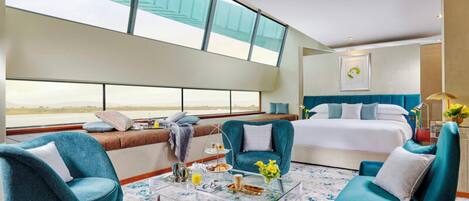 Habitación de hotel moderna con una cama grande, dos sillones azules, una mesa de centro de cristal y vistas al mar a través de amplias ventanas.