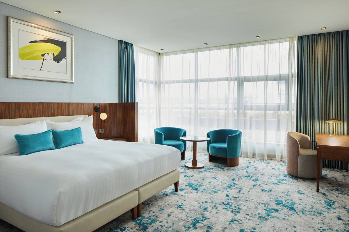Hotelkamer met een groot bed, twee blauwe fauteuils, een klein rond tafeltje en uitzicht op de stad door grote ramen.