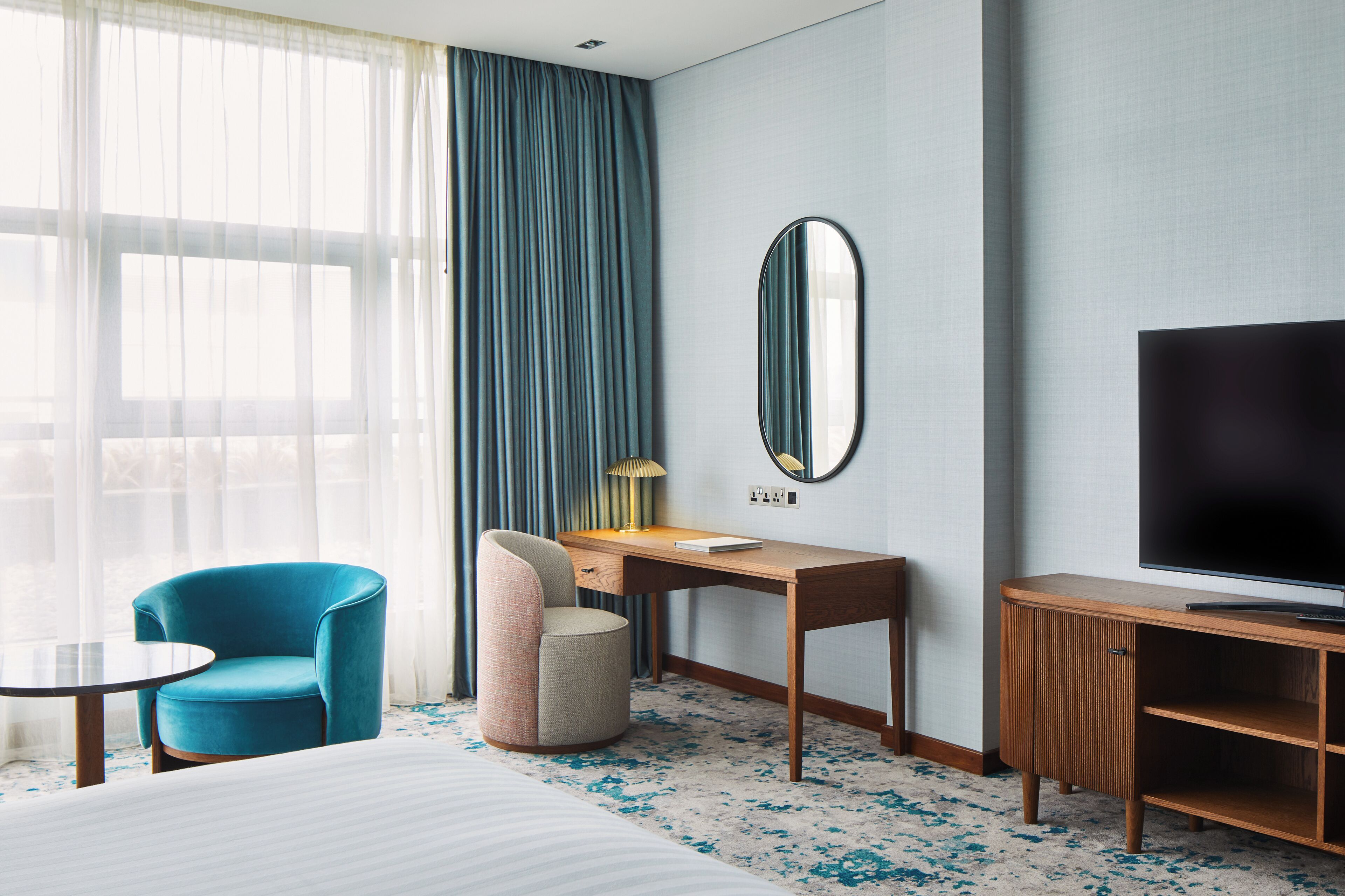 Habitación de hotel con cama, escritorio, silla, televisor y una ventana grande con cortinas.