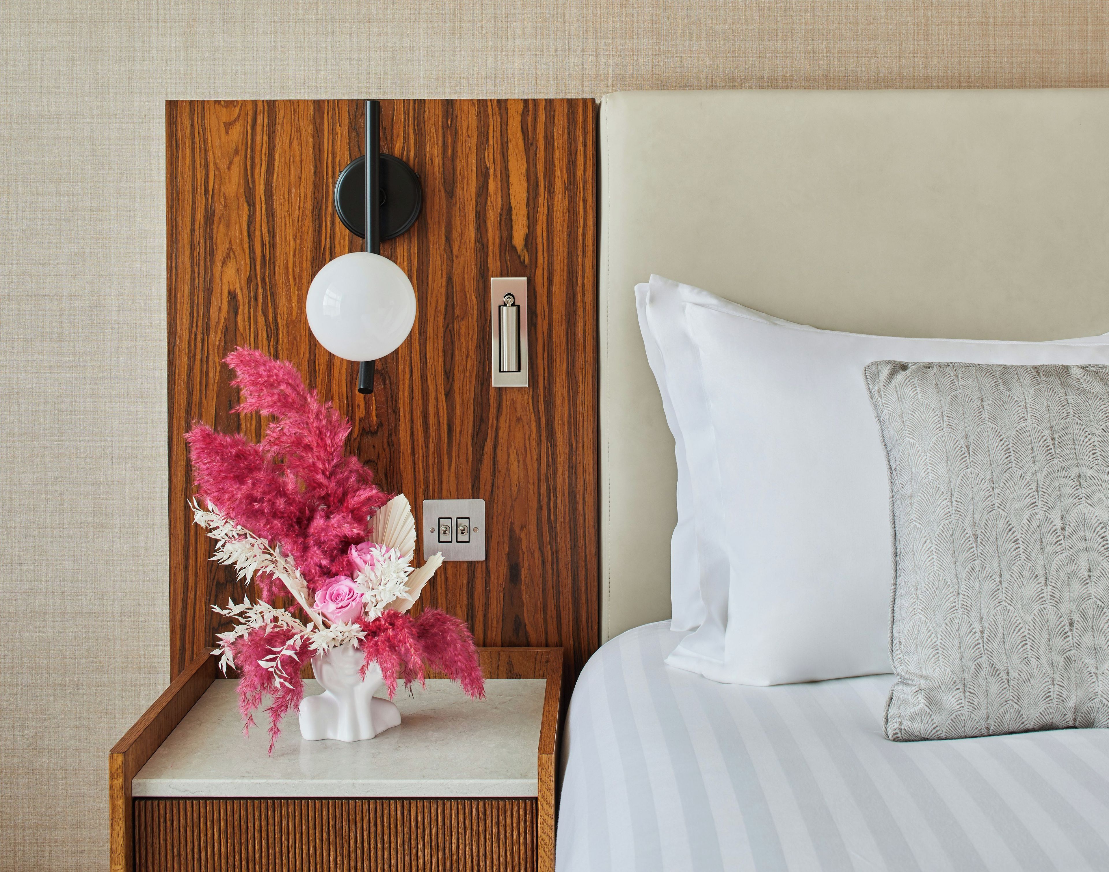 A neatly made hotel room with a wooden headboard, a bedside table with a lamp, a vase with pink flowers, and a bed with white and gray bedding.